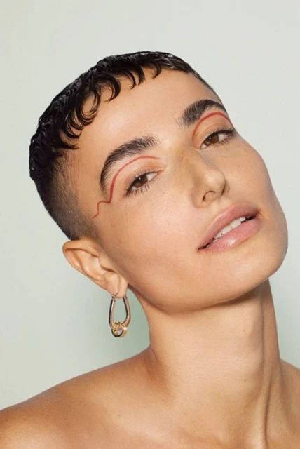 Close-up portrait of a woman with an asymmetric haircut, striking red eyeliner, gold hoop earrings, and a neutral background.