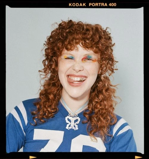 Young woman with curly hair, wearing a sports jersey and necklace, sticking her tongue out, with colorful makeup and a playful expression.