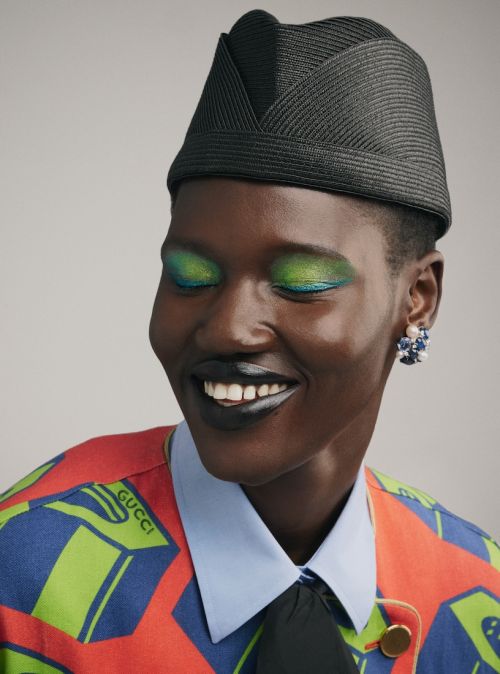 Smiling Black woman with green eye shadow and a black hat. The model is wearing a red, blue and green printed jacket and floral earrings.