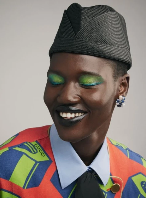 Smiling Black woman with green eye shadow and a black hat. The model is wearing a red, blue and green printed jacket and floral earrings.