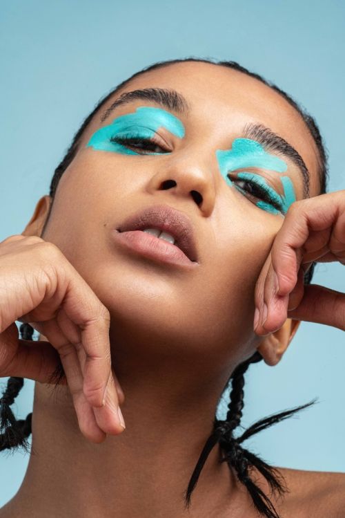 Close-up of a young adult woman with black braided hair, tan skin, and blue makeup on her eyelids, looking at the camera against a blue backdrop.