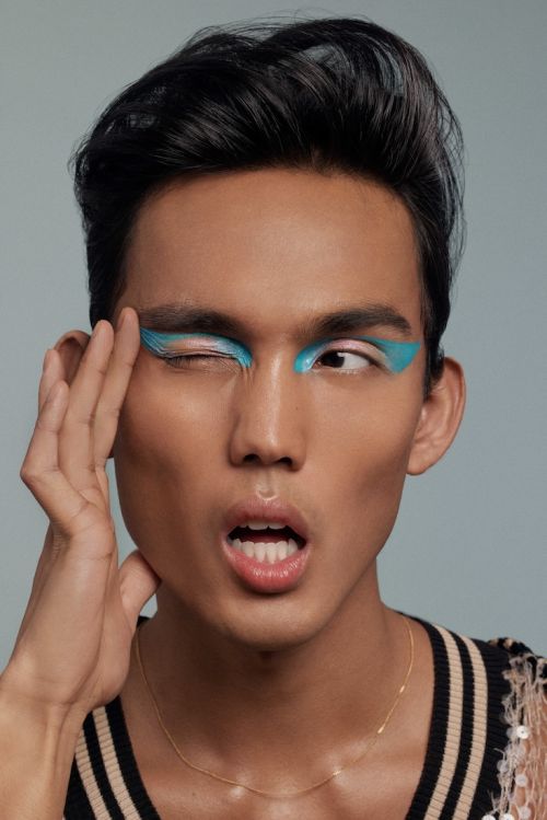 Close-up of an adult with vibrant turquoise eye makeup, striking a pose with a hand near their face, set against a grey background.