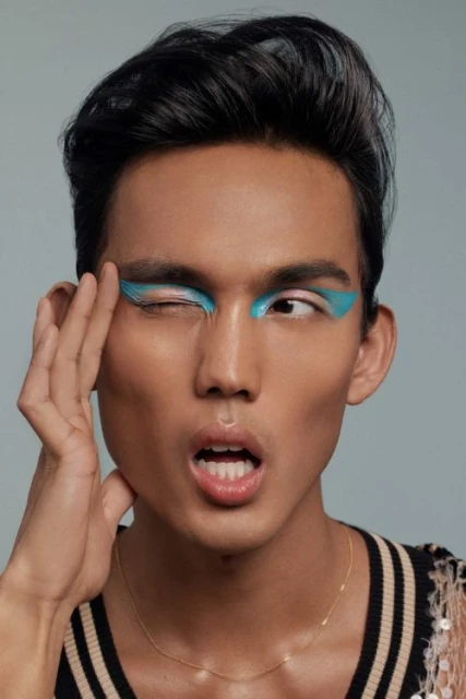 Close-up of an adult with vibrant turquoise eye makeup, striking a pose with a hand near their face, set against a grey background.