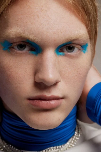 Close-up portrait of a young adult with blue feathered makeup around the eyes, a blue ruffled collar, and a rhinestone necklace.