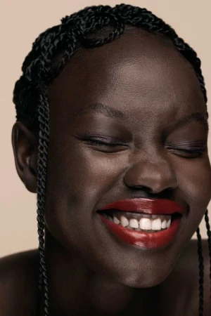 Close-up portrait of a smiling Black woman with braided hair and glossy red lipstick against a neutral beige background.