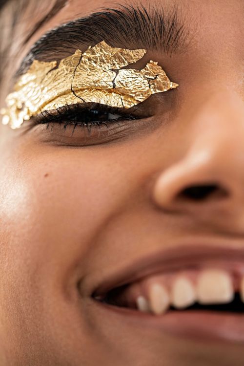 Close-up of a smiling adult female with golden foil eye makeup, fair skin, and an eye-level portrait orientation.