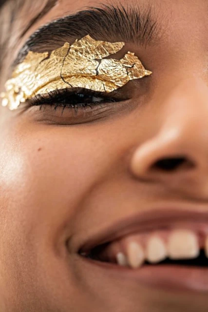 Close-up of a smiling adult female with golden foil eye makeup, fair skin, and an eye-level portrait orientation.