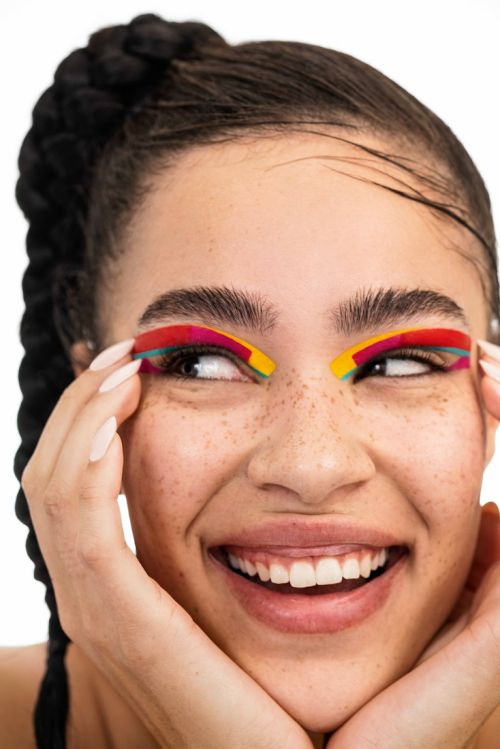 Close-up shot of a smiling woman with colorful makeup and braided hair posing against a white background, showcasing beauty and style.