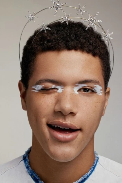 Close-up portrait of a smiling adult male with curly hair wearing a star crown and white face paint, against a neutral background.