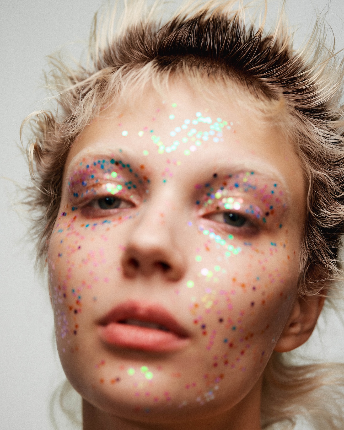 Close-up of a woman with light hair and glitter makeup around her eyes and on her face against a neutral background.