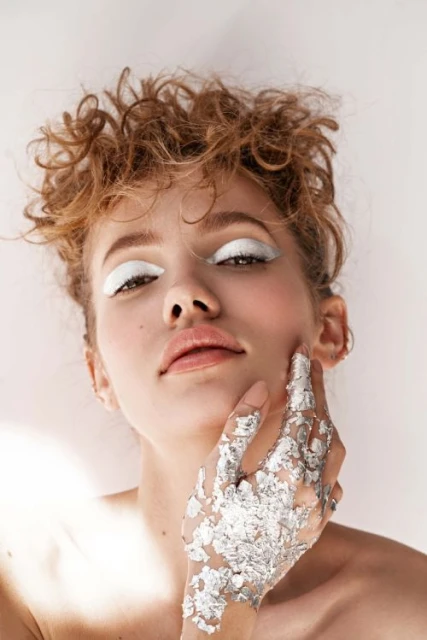 Close-up of a woman with light skin and curly hair, showcasing bold white eye makeup and a silver-covered hand against a plain background.