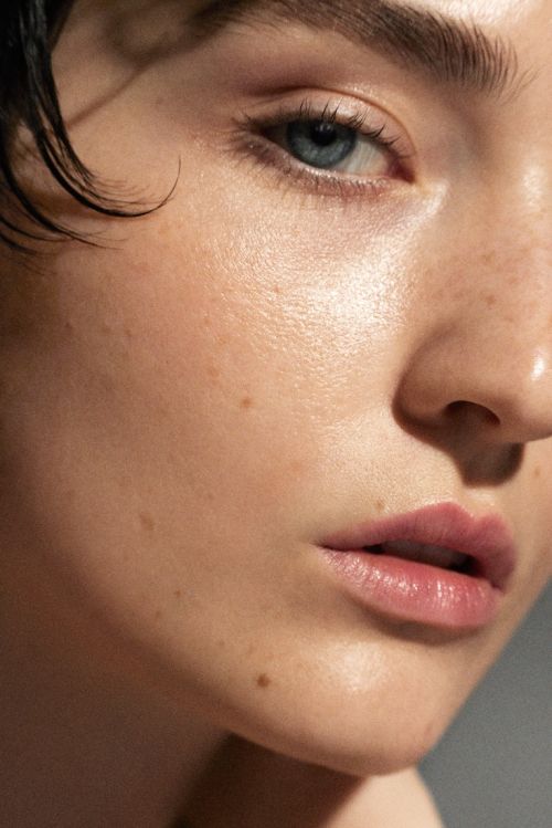 Close-up portrait of a young adult woman with fair skin, freckles, and blue eyes. Wet dark hair is visible. Beauty shot with natural lighting.