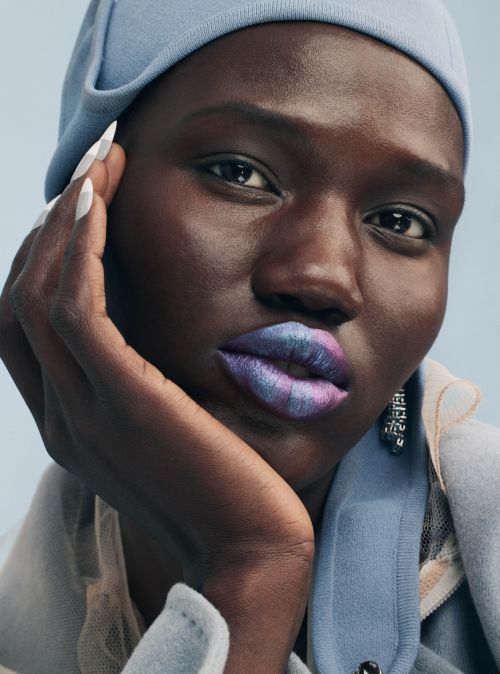 Close-up portrait of a Black woman with bright, multicolored lipstick and a light blue hat against a light blue background.