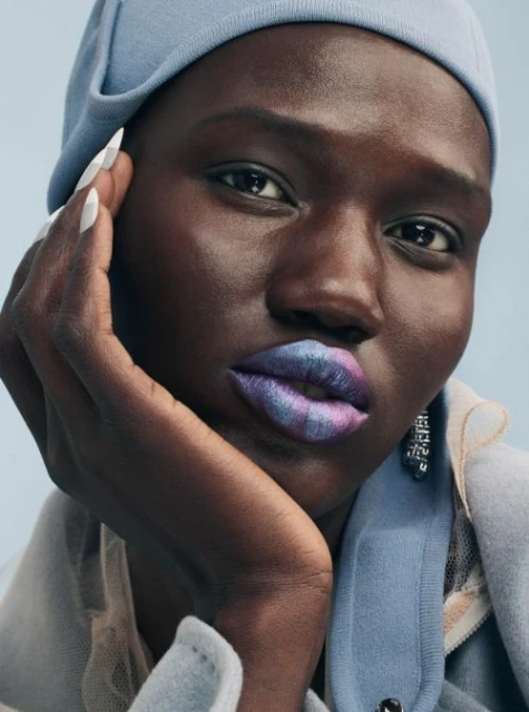 Close-up portrait of a Black woman with bright, multicolored lipstick and a light blue hat against a light blue background.