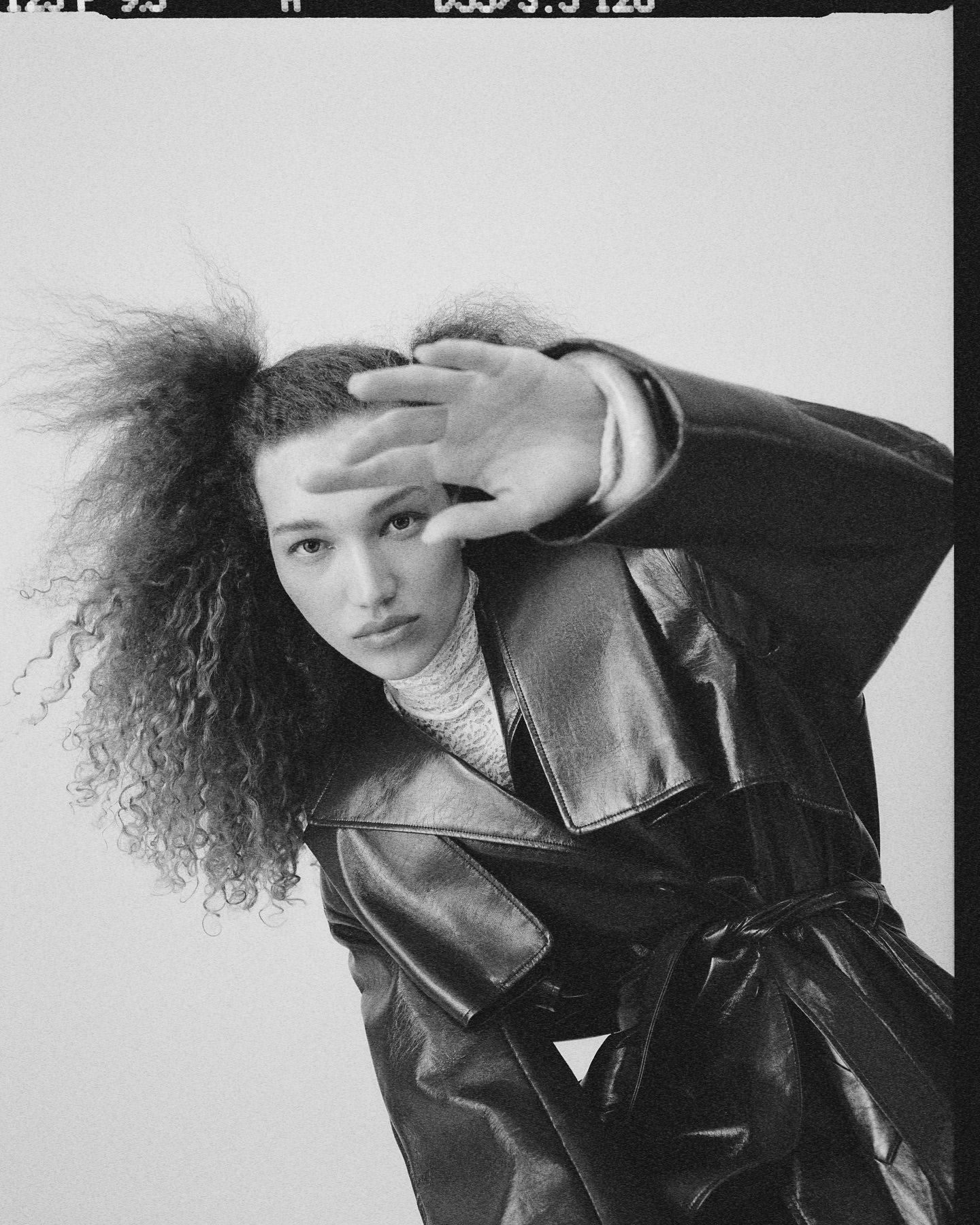 Portrait of a stylish young woman with curly hair wearing a black leather trench coat and a turtleneck in a studio setting.