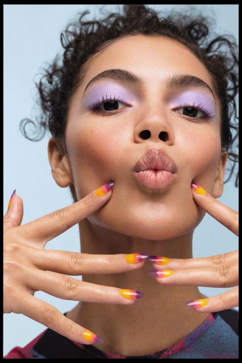 Close-up of a woman with colorful makeup. She has light purple eyeshadow and orange and purple nail polish on her nails.