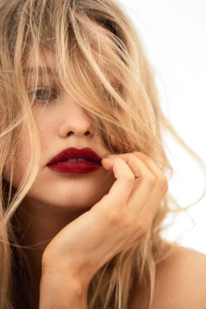Close-up portrait of a woman with blonde hair across her face, deep red lipstick, and hand near her chin against a white background.