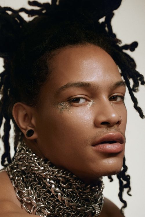 Close-up portrait of a young adult male with locs, wearing a metal chain necklace, glitter eye makeup, and against a neutral backdrop.