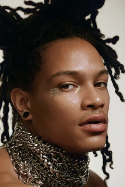 Close-up portrait of a young adult male with locs, wearing a metal chain necklace, glitter eye makeup, and against a neutral backdrop.
