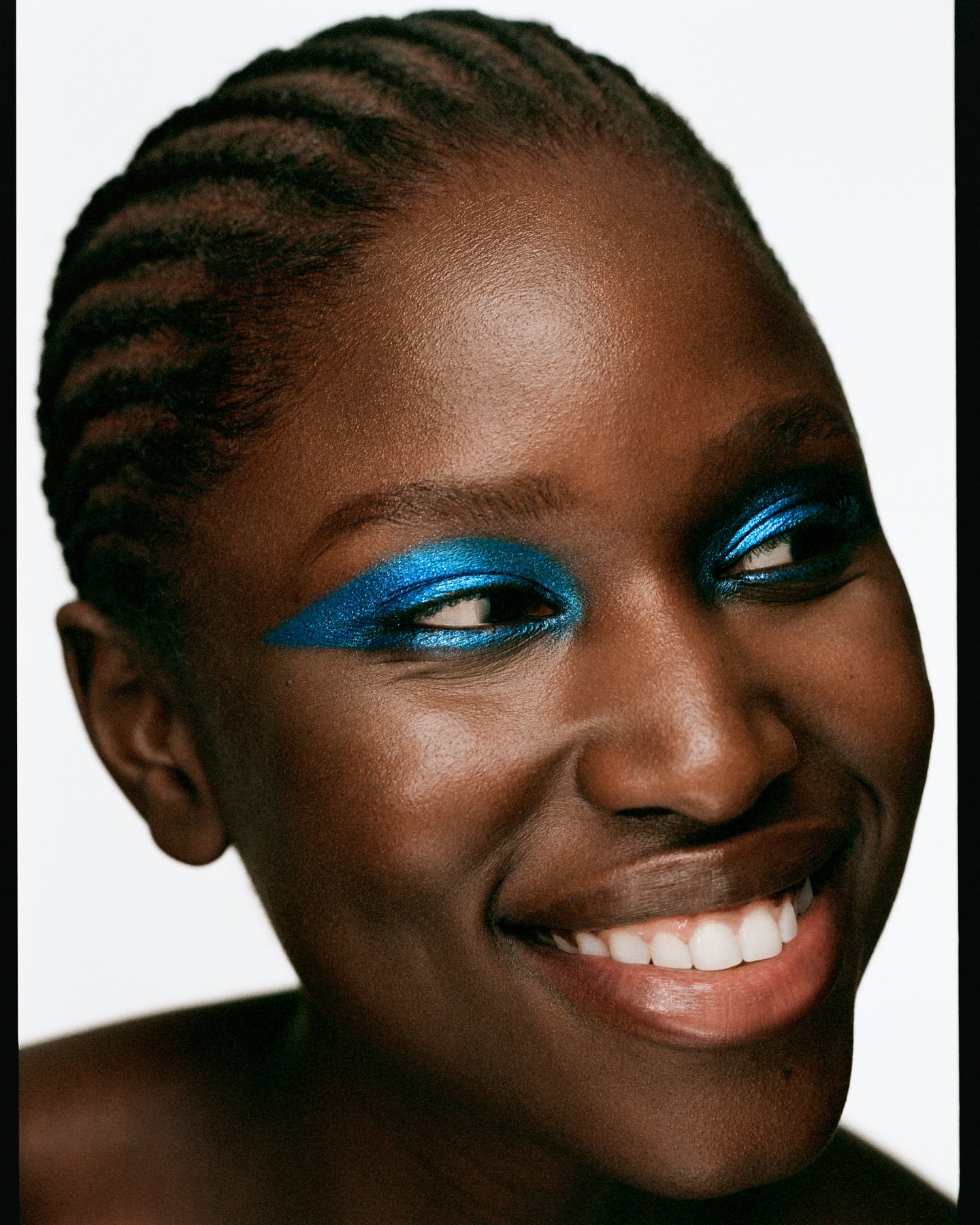 Close-up shot of a smiling African-American woman with bright blue eyeshadow and braided hair against a white background.