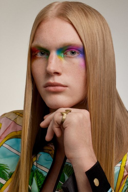 Close-up of a young adult with blond hair and rainbow eye makeup wearing a tropical-patterned shirt and statement ring against white backdrop.