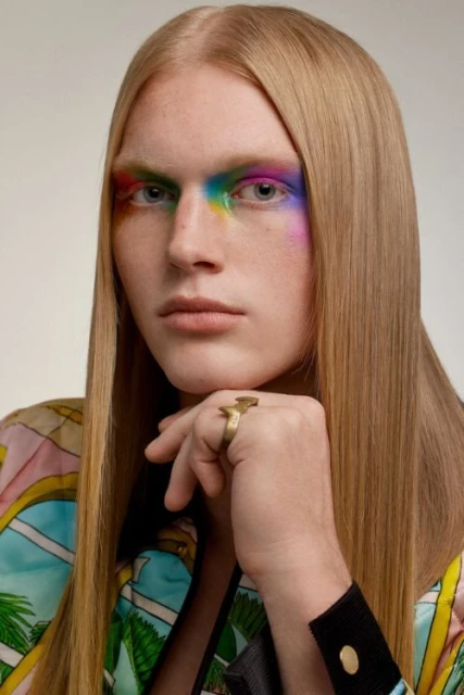 Close-up of a young adult with blond hair and rainbow eye makeup wearing a tropical-patterned shirt and statement ring against white backdrop.