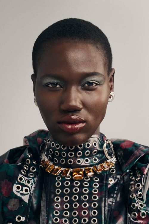 Close-up studio shot of a Black woman with short hair wearing a high-fashion jewel necklace and vinyl-style jacket with grommets.
