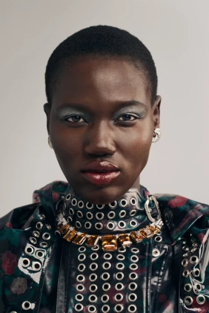 Close-up studio shot of a Black woman with short hair wearing a high-fashion jewel necklace and vinyl-style jacket with grommets.