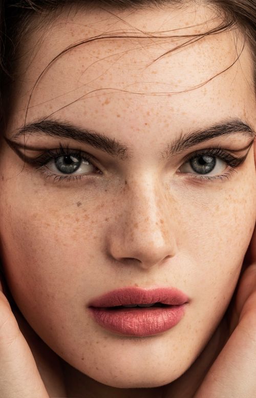 Close-up portrait of a woman with fair skin and freckles. She has a minimalist makeup look featuring winged eyeliner and mauve lipstick.