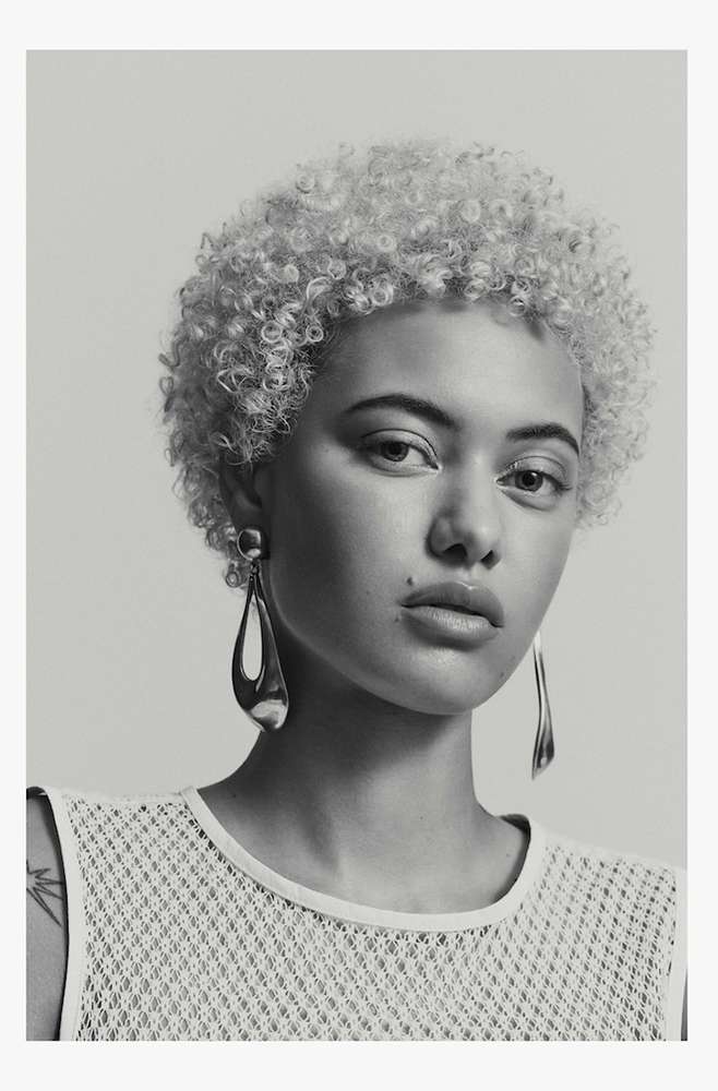 Close-up portrait of a light-skinned woman with curly blonde hair wearing teardrop earrings and a net-textured top against a grey backdrop.