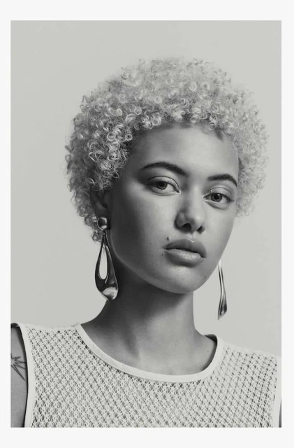 Close-up portrait of a light-skinned woman with curly blonde hair wearing teardrop earrings and a net-textured top against a grey backdrop.