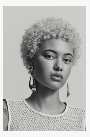 Close-up portrait of a light-skinned woman with curly blonde hair wearing teardrop earrings and a net-textured top against a grey backdrop.