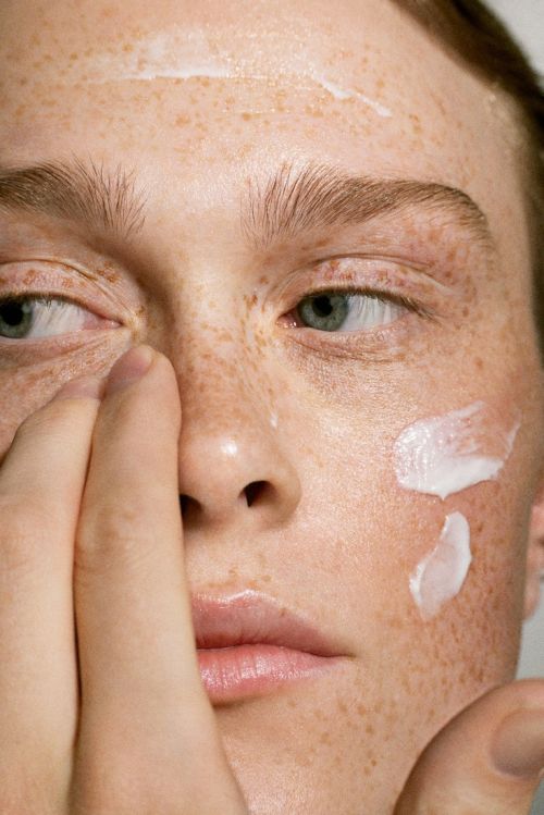 Close-up of a fair-skinned young adult female applying white cream to her face, showing skin texture and freckles, with a neutral expression.