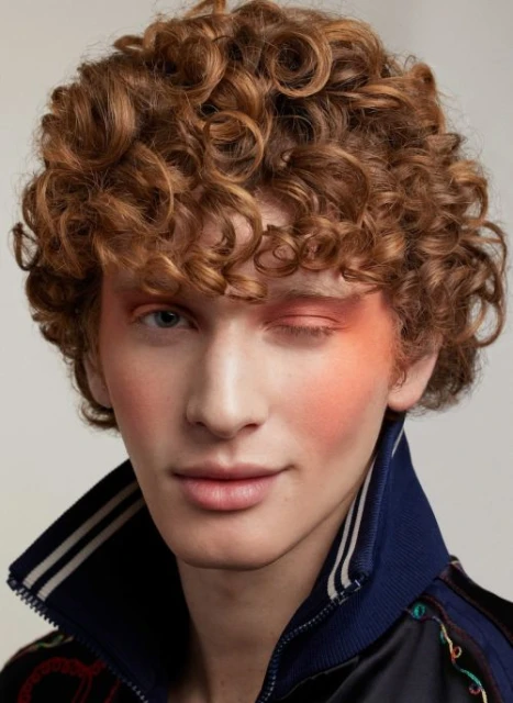 Close-up studio portrait of a young adult man with curly auburn hair, wearing dark jacket and colorful eye makeup with a slight smile.