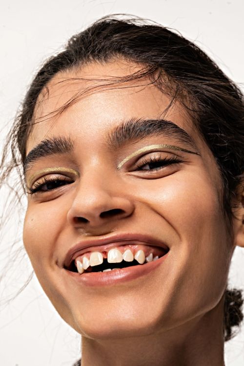 Close-up studio shot of a smiling woman with a gap in her teeth, wearing gold eyeliner, against a neutral background.