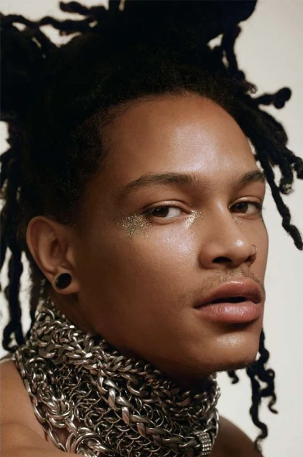 Close-up portrait of a person with locs, wearing glitter around the eyes, and a heavy silver chain necklace, against a white background.