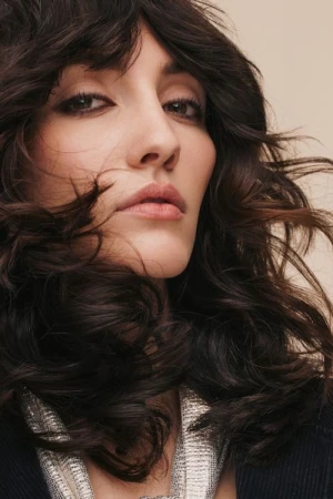 Close-up portrait of a woman with dark layered wavy hair and textured silver top against a neutral background.