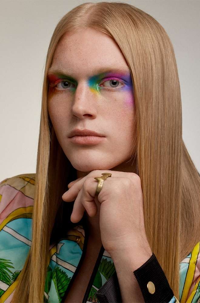 Close-up portrait of a fair-skinned adult with straight blonde hair, rainbow makeup on the eyes, and a gold ring on the hand.