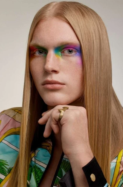 Close-up portrait of a fair-skinned adult with straight blonde hair, rainbow makeup on the eyes, and a gold ring on the hand.