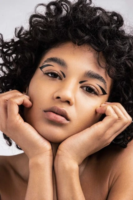 Close-up beauty portrait of a curly-haired woman with graphic black winged eyeliner against a pale gray backdrop in a photo studio.