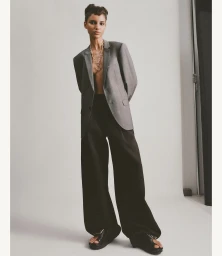 Model wearing a grey blazer, black trousers, and a statement chain necklace against a light grey studio background.