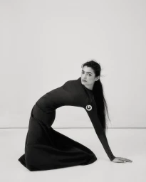 Monochrome portrait of a woman arching her back wearing a dark long-sleeve dress and spiral pendant against a seamless white background.