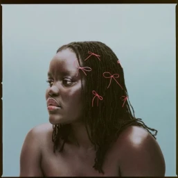 Portrait of a Black woman with dark braided hair adorned with small red bows against a solid greyish-blue background.