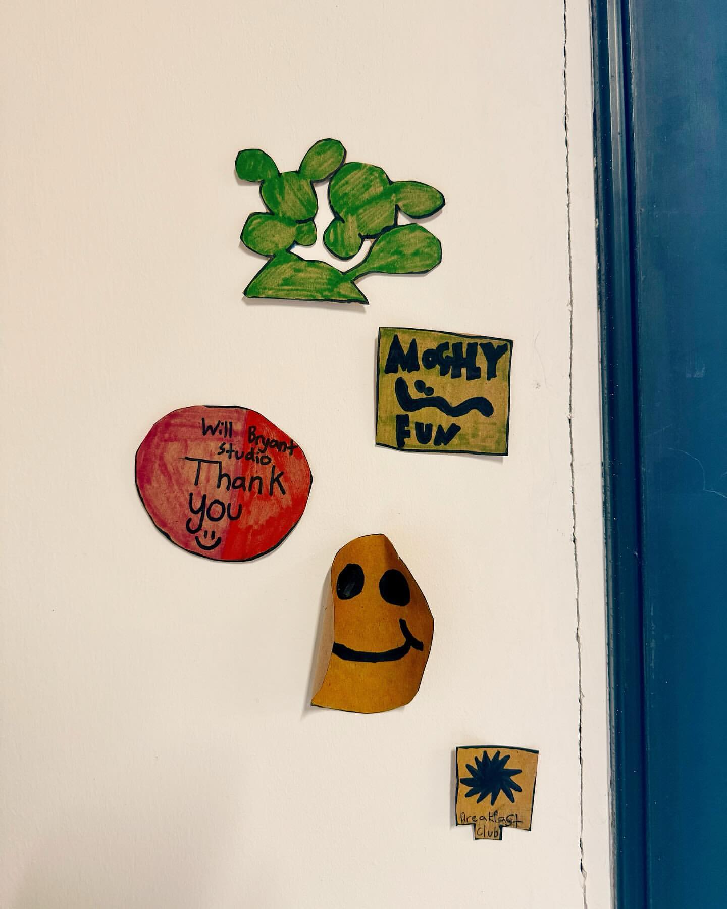 A collection of colorful handmade paper art and drawings, including cartoon faces and a green cactus, affixed to a white surface.