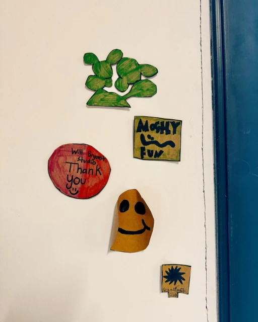 A collection of colorful handmade paper art and drawings, including cartoon faces and a green cactus, affixed to a white surface.