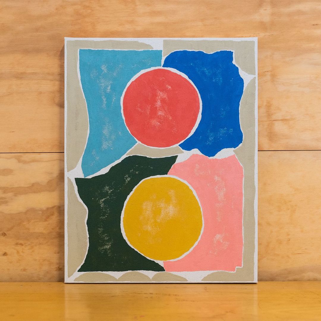 Abstract painting featuring overlapping geometric shapes in pink, blue, green, yellow, and red on a textured canvas against a wood background.