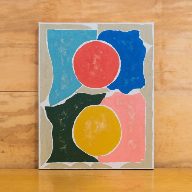Abstract painting featuring overlapping geometric shapes in pink, blue, green, yellow, and red on a textured canvas against a wood background.