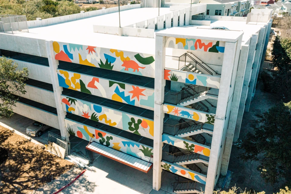 Exterior view of a multi-story parking garage with colorful abstract mural art, concrete walls, and open staircases.