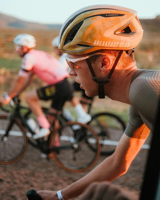 A cyclist wearing a helmet rides on a road, focusing on the man in the foreground, with other cyclists and blurred background.