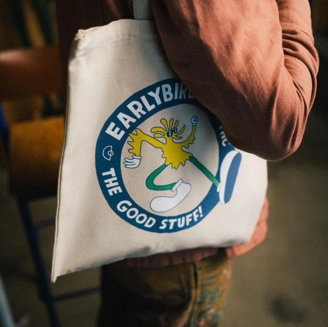 Canvas tote bag featuring cartoon graphic of a yellow and green figure, held by a person in a terracotta shirt, indoors.